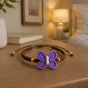 May include: A handmade bracelet featuring a purple crochet butterfly charm. The bracelet has a brown cord with gold-colored beads. The butterfly has white accents. The bracelet is displayed on a wooden surface.