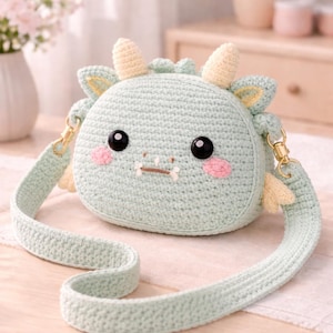 May include: A light green crocheted round purse shaped like a dragon with black eyes, pink cheeks, and yellow horns and wings. The purse has a matching strap and gold-colored hardware. The background includes a table and flowers.