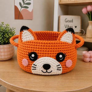 May include: An orange crocheted fox basket with black and white features. The basket has a white muzzle, black eyes, and pink cheeks. It has two handles and fox ears. The basket is displayed on a wooden table.