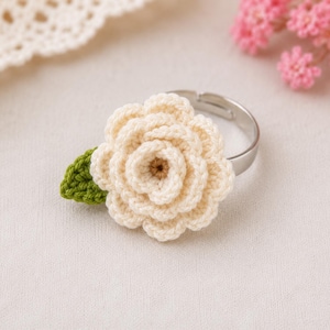 Puede incluir: Anillo de ganchillo hecho a mano con una flor color crema, centro marrón y una hoja verde. La flor está unida a un anillo ajustable plateado. El anillo se encuentra sobre un fondo suave blanquecino.