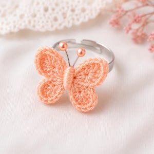 Puede incluir: Anillo de mariposa de ganchillo color melocotón, con una banda ajustable plateada. La mariposa tiene alas detalladas y antenas con pequeñas cuentas redondas. El anillo se presenta sobre un fondo blanco suave, sugiriendo un accesorio delicado y hecho a mano.