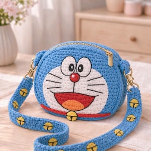 May include: A blue crochet crossbody bag featuring the cartoon character Doraemon. The bag has a gold zipper, a red mouth, and a yellow bell. The strap is blue with yellow bell accents.