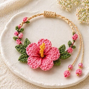 Può includere: Un braccialetto rosa all'uncinetto fatto a mano con un grande fiore di ibisco con stami gialli, circondato da foglie verdi e boccioli rosa. Il braccialetto ha un cordoncino beige intrecciato e dettagli di perline rosa.