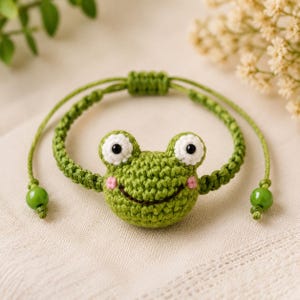 Peut inclure: Un bracelet en crochet vert représentant une tête de grenouille avec des yeux blancs et noirs, des joues roses et un sourire brodé. Le bracelet est composé d'un cordon tressé vert avec un nœud coulissant et une perle verte à chaque extrémité.