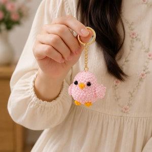 May include: A small, round, pink beaded chick keychain featuring a gold-toned metal ring and chain. The chick has two black bead eyes, a small yellow bead beak, and two yellow bead feet, all constructed from tiny, uniform pink beads.