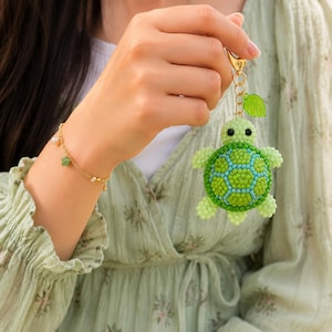 May include: A beaded turtle keyring featuring a light green and dark green shell with black bead eyes, attached to a gold-tone metal chain with a small green leaf charm. A delicate gold-tone chain bracelet with small white pearls and a green star charm.