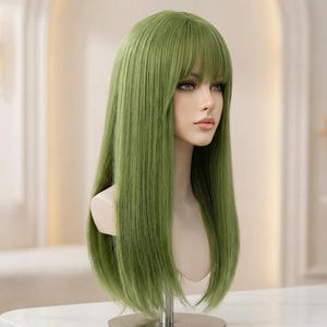 May include: A long, green wig with bangs is displayed on a mannequin head. The wig has a natural-looking style and is a vibrant shade of green. The mannequin is on a metal stand, set against a neutral background.