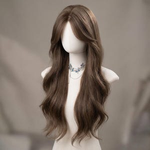 Op de afbeelding: Een lange, golvende bruine pruik met een zijscheiding, gepresenteerd op een mannequinhoofd. De pruik heeft een natuurlijke uitstraling met zachte lagen. Een zilveren ketting met blauwe vlinderaccenten siert de nek van de mannequin.