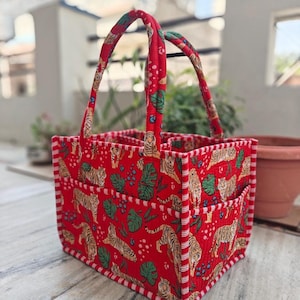 Peut inclure: Un panier en tissu matelassé rectangulaire avec un fond rouge vif orné d'un motif répétitif de tigres, de feuilles tropicales vertes et de petites baies bleues. Le sac possède deux anses robustes et des bordures rayées rouge et blanc.