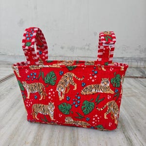May include: A red fabric tote bag with a repeating pattern of tigers, green leaves, and blue dots. The bag has two red and white striped handles with a velcro closure. The bag is designed for carrying items.