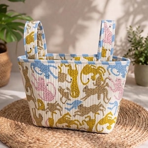 May include: A quilted fabric tote bag featuring a repeating pattern of stylised leopards in yellow, pink, blue, and tan on an off-white background. The bag has two upright handles and a light blue and white striped trim along the top edge.