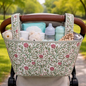 May include: A fabric stroller organizer featuring a floral pattern with pink flowers and green vines on a cream background. The organizer hangs from a brown leather-covered handlebar and contains a light blue pouch, two baby bottles, and a teddy bear.