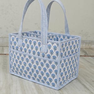 Puede incluir: Un organizador de almacenamiento rectangular de tela con un estampado floral en azul claro y blanco. El organizador cuenta con dos asas verticales y bolsillos exteriores, todo ribeteado con tela a rayas azul claro y blanco.
