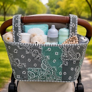 May include: A quilted stroller organizer featuring a dark gray and teal paisley bandana pattern with black and white striped trim. The bag contains a light blue pouch, white rolled cloths, a small plush bear, two baby bottles, and wooden beads.