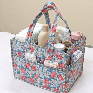 Puede incluir: Una cesta de almacenamiento rectangular de tela acolchada con un estampado floral azul y rosa y un borde a cuadros grises y blancos. La cesta tiene dos asas y varios bolsillos exteriores que contienen artículos de bebé.