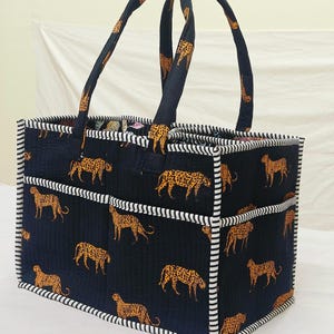 May include: A rectangular quilted tote bag featuring a dark navy blue fabric with a repeating orange and black leopard print pattern. The bag includes two matching fabric handles and is finished with white and black striped fabric trim.