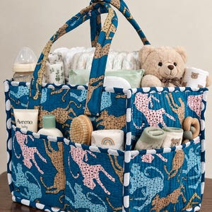 Peut inclure: Un panier de rangement rectangulaire en tissu avec deux anses, présentant un fond bleu sarcelle avec un motif répétitif de guépards beige, rose et bleu clair. Les bords sont ornés de pois blancs et bleus, et il contient des articles de bébé.