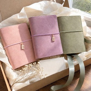 May include: Three rectangular notebooks in soft pink, mauve, and olive green, each featuring a smooth texture and a thin elastic band closure. Two notebooks are adorned with a small, gold-toned letter E charm embellished with white faux pearls.