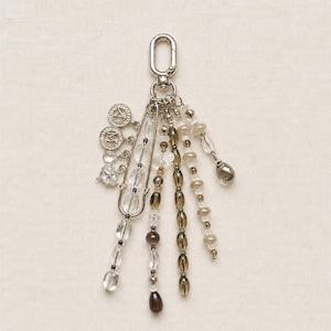 May include: A silver-toned metal keychain featuring a lobster clasp, a large safety pin, and multiple dangling strands of clear, translucent, and dark brown beads. Two small circular charms with the letters A and M are attached to the safety pin.
