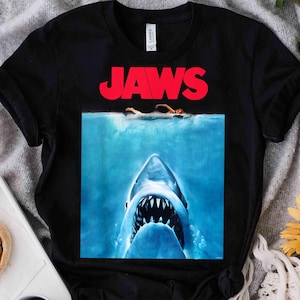 Op de afbeelding: Een zwart t-shirt met korte mouwen en een grafische print van de Jaws-filmposter. Het ontwerp toont de rode tekst JAWS boven een illustratie van een zwemmer aan het wateroppervlak met een grote haai die van onderaf nadert.