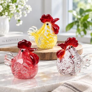 May include: Three glass rooster figurines featuring a red, yellow, and white speckled design. Each rooster has a bright red comb and wattle, a clear glass tail, and a rounded body. The figurines are crafted with a textured, speckled glass finish.