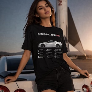 Puede incluir: Camiseta negra con el texto "NISSAN GT-R" y un gráfico de un coche deportivo blanco con especificaciones. Una persona se apoya en un coche blanco. Un cohete está en el fondo.