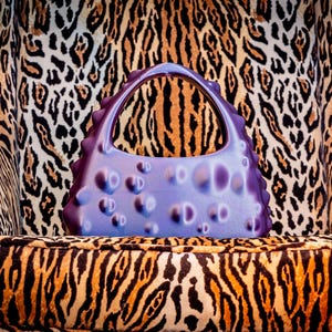 Puede incluir: Un bolso lavanda con un diseño texturizado único con protuberancias redondeadas. El bolso tiene un asa curva y está sobre una silla con un llamativo estampado animal. La estética general es moderna y llamativa.