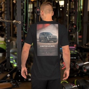 Puede incluir: Camiseta gris oscuro con un gráfico de Land Rover Defender en la espalda. El gráfico muestra un SUV gris en un paisaje desértico, con la palabra "DEFENDER" debajo. La camiseta es de manga corta y tiene cuello redondo.