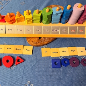 May include: A wooden educational toy with colorful geometric shapes and pegs. The toy has a wooden base with the numbers one through ten printed on it. There are also matching number cards and shapes. The colors include red, orange, yellow, green, blue, and purple.