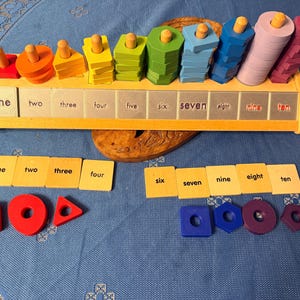 May include: A wooden educational toy with colorful geometric shapes and pegs. The toy has a wooden base with the numbers one through ten printed on it. There are also matching number cards and shapes. The colors include red, orange, yellow, green, blue, and purple.
