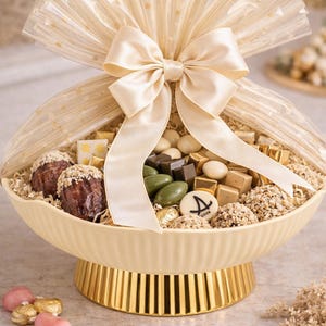 May include: A decorative gift basket filled with assorted chocolates and candies. The basket is cream-colored with a gold base and a large cream-colored bow. The chocolates include truffles, wrapped candies, and chocolate-covered nuts.