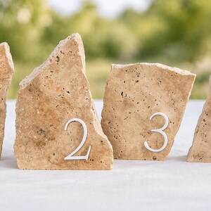 May include: Four beige stone table number holders, each with a white number from one to four. The stones have a rough, textured surface and are arranged on a white surface with a blurred green background.
