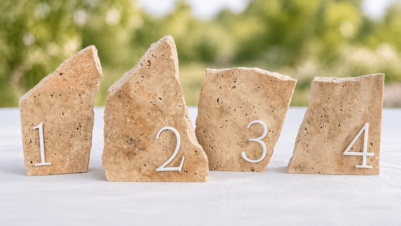 May include: Four beige stone table number holders, each with a white number from one to four. The stones have a rough, textured surface and are arranged on a white surface with a blurred green background.