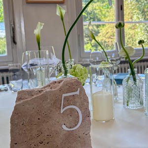 May include: A natural stone table number five with a white numeral, set on a white tablecloth. The table is decorated with clear glass vases, white flowers, and candles, creating an elegant ambiance.
