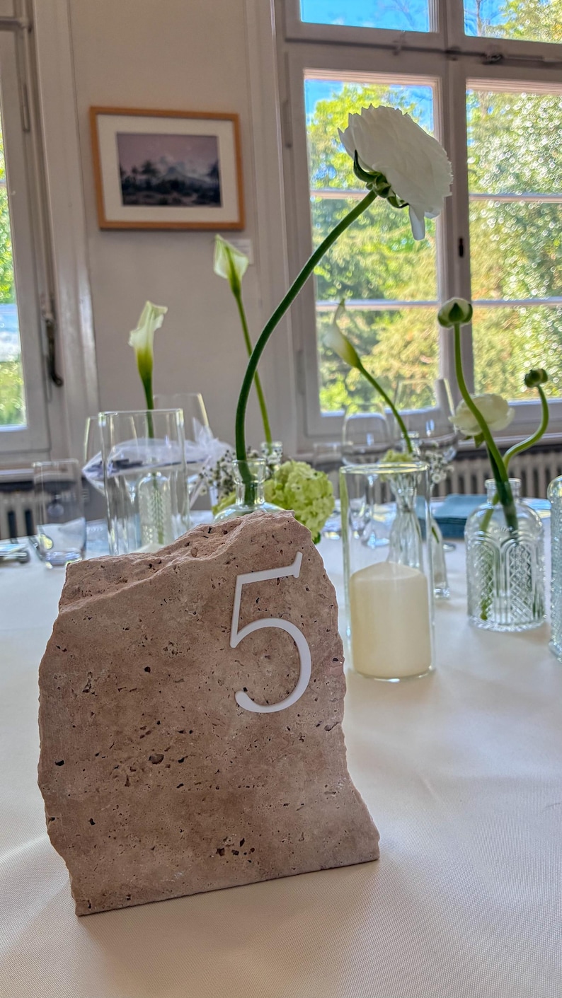 May include: A natural stone table number five with a white numeral, set on a white tablecloth. The table is decorated with clear glass vases, white flowers, and candles, creating an elegant ambiance.