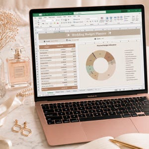 May include: A rose gold MacBook Air laptop displaying a wedding budget planner spreadsheet. The screen shows a pie chart and budget categories. A bottle of Coco Chanel perfume, gold earrings, and a notebook with a pen are also visible.