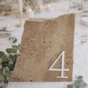 May include: A natural stone table number 4, perfect for weddings or events. The beige stone has a rough texture and a white number 4 attached to the front. Greenery is placed around the stone.