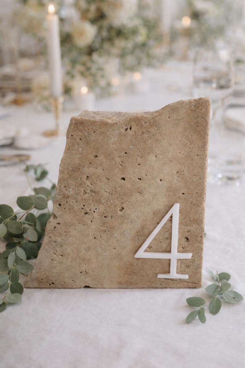 May include: A natural stone table number 4, perfect for weddings or events. The beige stone has a rough texture and a white number 4 attached to the front. Greenery is placed around the stone.