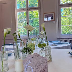 May include: A table setting with white tablecloth, floral arrangements, and a stone table number. The flowers include white calla lilies, roses, and small green blooms in clear glass vases. A large white candle is also present.