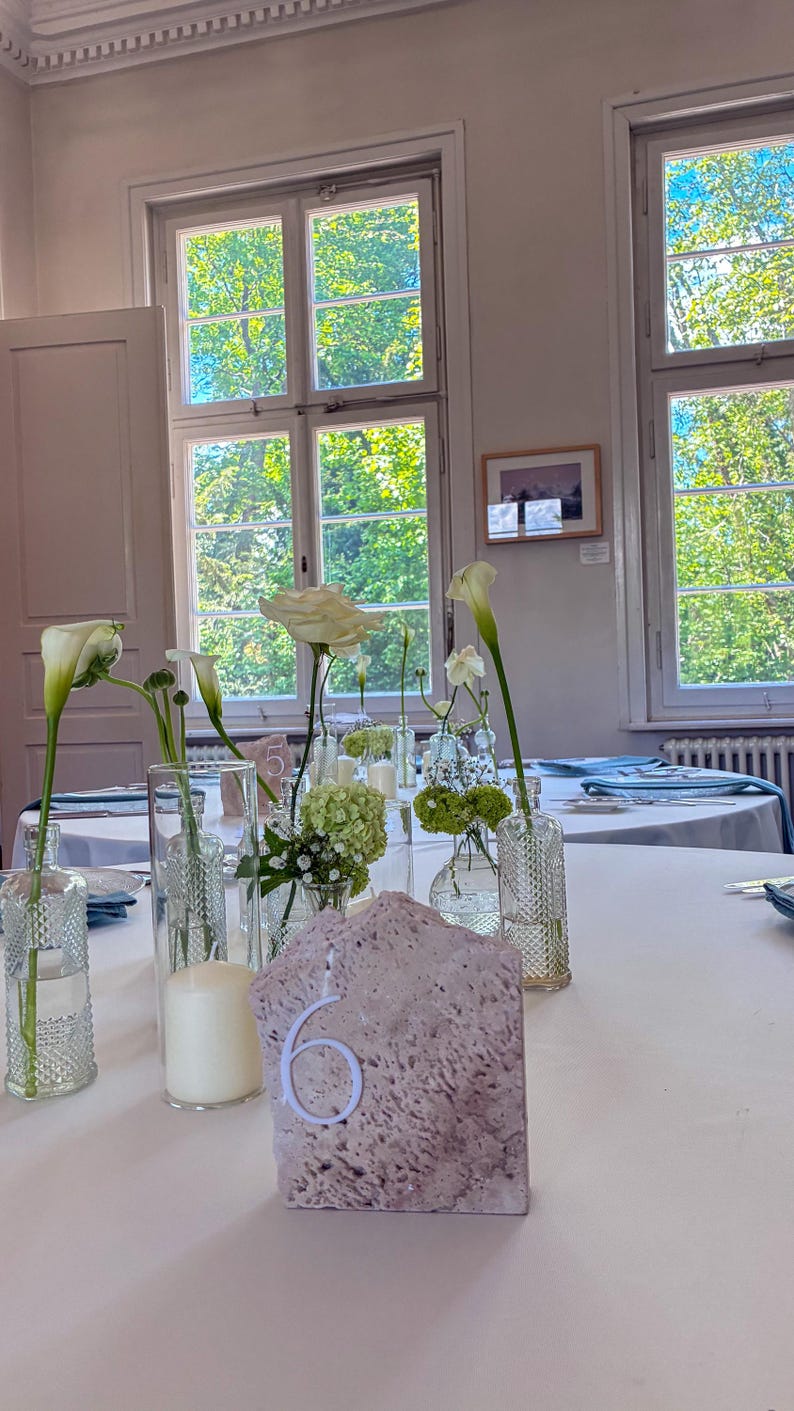 May include: A table setting with white tablecloth, floral arrangements, and a stone table number. The flowers include white calla lilies, roses, and small green blooms in clear glass vases. A large white candle is also present.