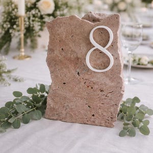 May include: A rustic, rectangular stone table number eight. The number is white and affixed to the front of the stone. The stone is a light brown color and has a rough, textured surface. The table is set for a wedding.