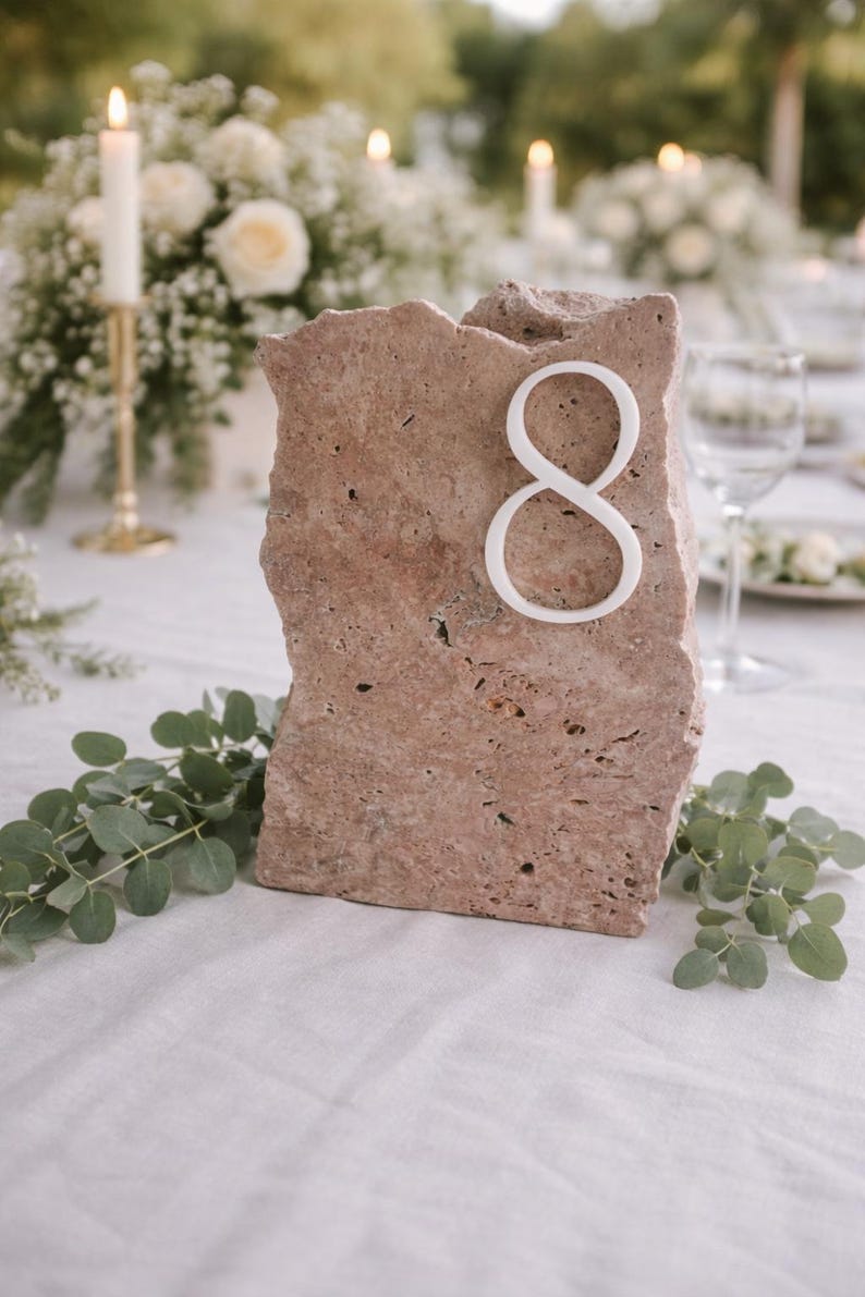 May include: A rustic, rectangular stone table number eight. The number is white and affixed to the front of the stone. The stone is a light brown color and has a rough, textured surface. The table is set for a wedding.
