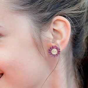 May include: A circular stud earring featuring a central clear stone surrounded by a ring of small, round, deep pink gemstones. The design resembles a flower with petals arranged in a radial pattern, set in a metallic frame against an earlobe.