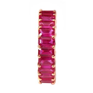 May include: An eternity band ring featuring a continuous row of vibrant, rectangular emerald-cut pink stones set in a polished rose gold metal frame. The stones are secured by small, rounded prongs that hold each gem in place along the band.
