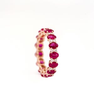 May include: An eternity band ring crafted from rose gold, featuring a repeating pattern of oval-cut vibrant red rubies alternating with small, round, clear white diamonds set in a continuous circular design around the entire band.