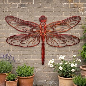 May include: A large, flat, metal dragonfly wall sculpture with a reddish-brown, gradient finish. The piece features intricate, cut-out wing patterns and a segmented body, mounted centrally on a grey brick wall above several potted plants.