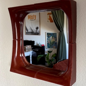 Puede incluir: Un espejo de pared cuadrado con un marco grueso, brillante y de color marrón rojizo oscuro con esquinas interiores redondeadas. El marco está construido con cuatro segmentos distintos que se unen en el centro de cada lado, creando un borde curvo.