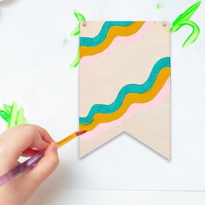May include: A wooden banner with a wavy design in teal, gold, and pink paint. The banner has a natural wood grain background and is rectangular with a pointed bottom. The banner is hanging on a white surface.