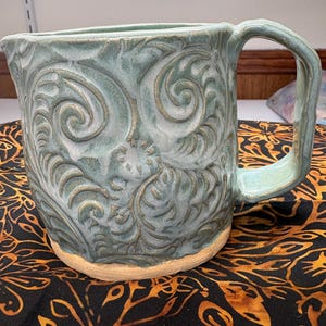 Puede incluir: Jarra de cerámica artesanal con esmalte azul claro y intrincados patrones florales en relieve. La jarra tiene un asa resistente y una base natural sin esmaltar. El diseño presenta detalles en espiral y una estética rústica.