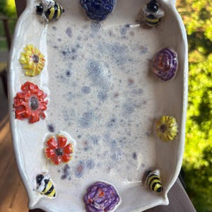 May include: A rectangular ceramic tray with a speckled off-white glaze, featuring three-dimensional decorative elements along the inner rim. These include two yellow and black bees, two purple flowers, two yellow flowers, and two orange flowers.